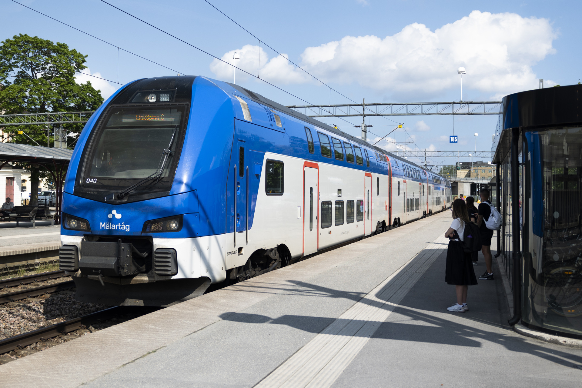 A blue and white Mälartåg train is at a platform on a sunny day. Some passengers are waiting on the platform, and the sky is clear with a few clouds.