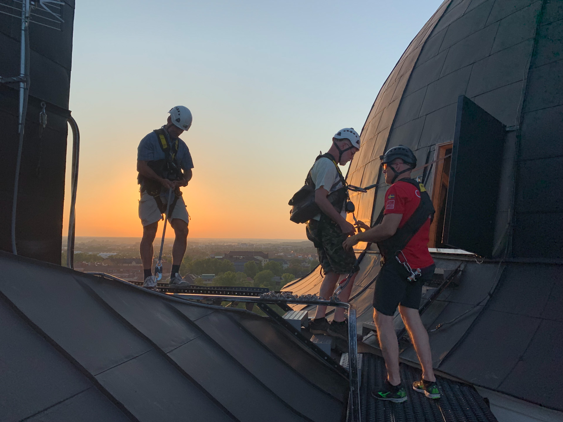 Tre personer iförda klätterutrustning står på ett tak vid solnedgången. Två av dem förbereder sig för en aktivitet medan den tredje håller i säkerhetslinor.
