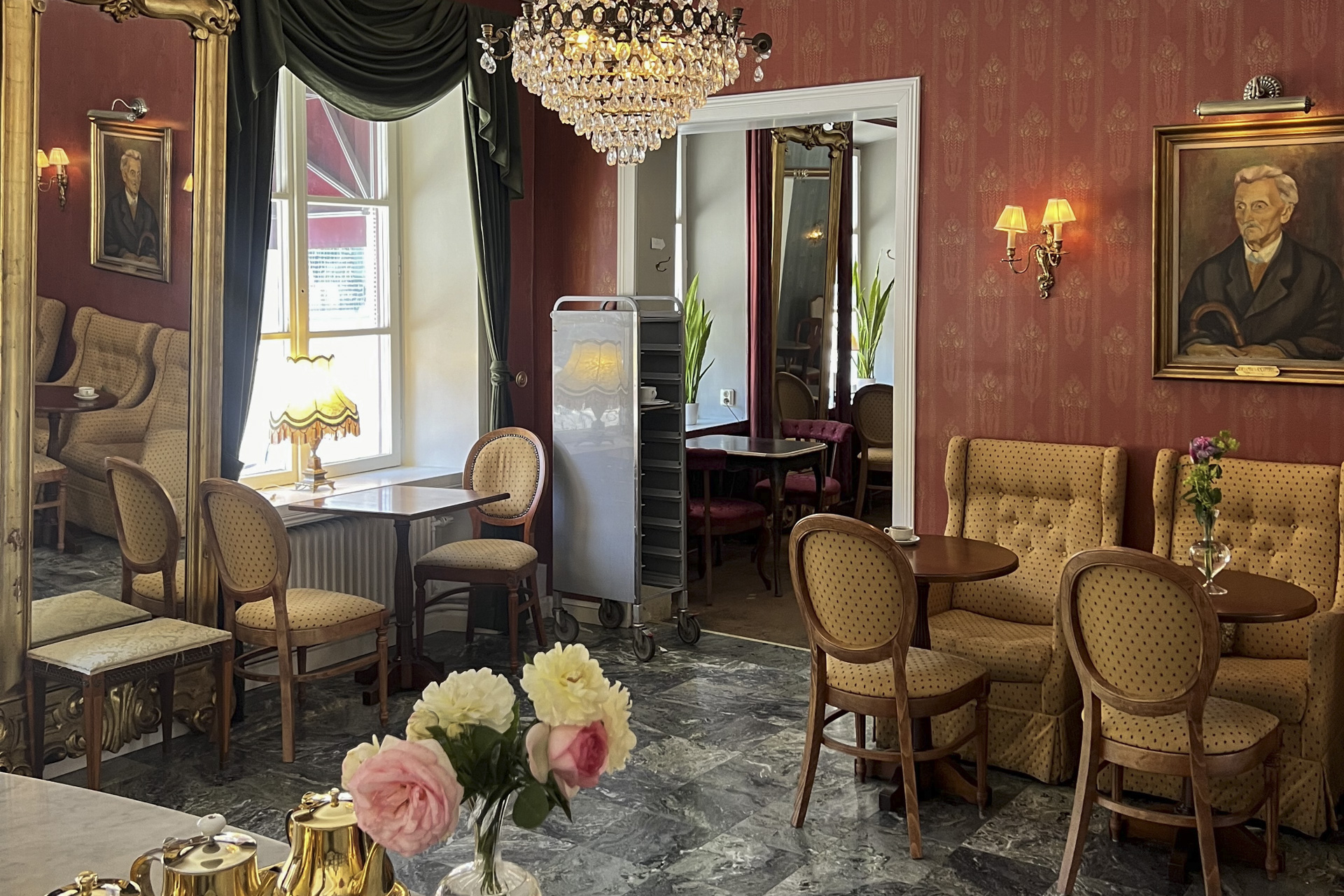 Ett elegant café med röda tapeter, kristallkrona, speglar, klassiska porträtt och stoppade stolar vid små runda bord.