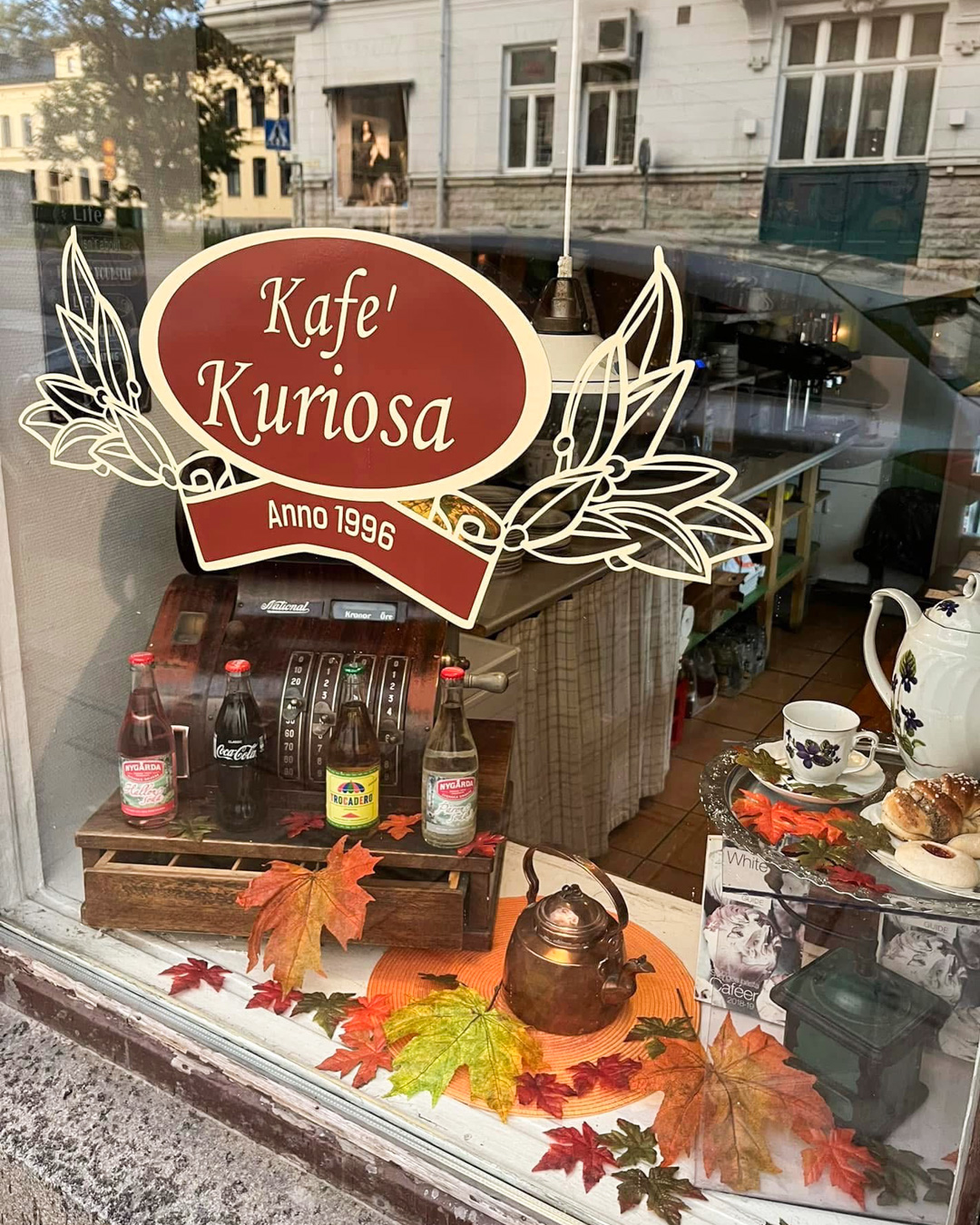 Ett skyltfönster med texten ”Kafé Kuriosa Anno 1996”, gamla flaskor, en kaffekanna och höstlöv som dekoration.
