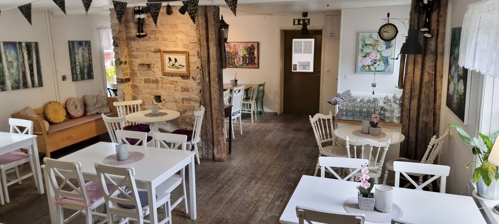 Ett mysigt café med vitmålade träbord och stolar, rustik tegelvägg, färgglada kuddar på en bänk och tavlor på väggarna.