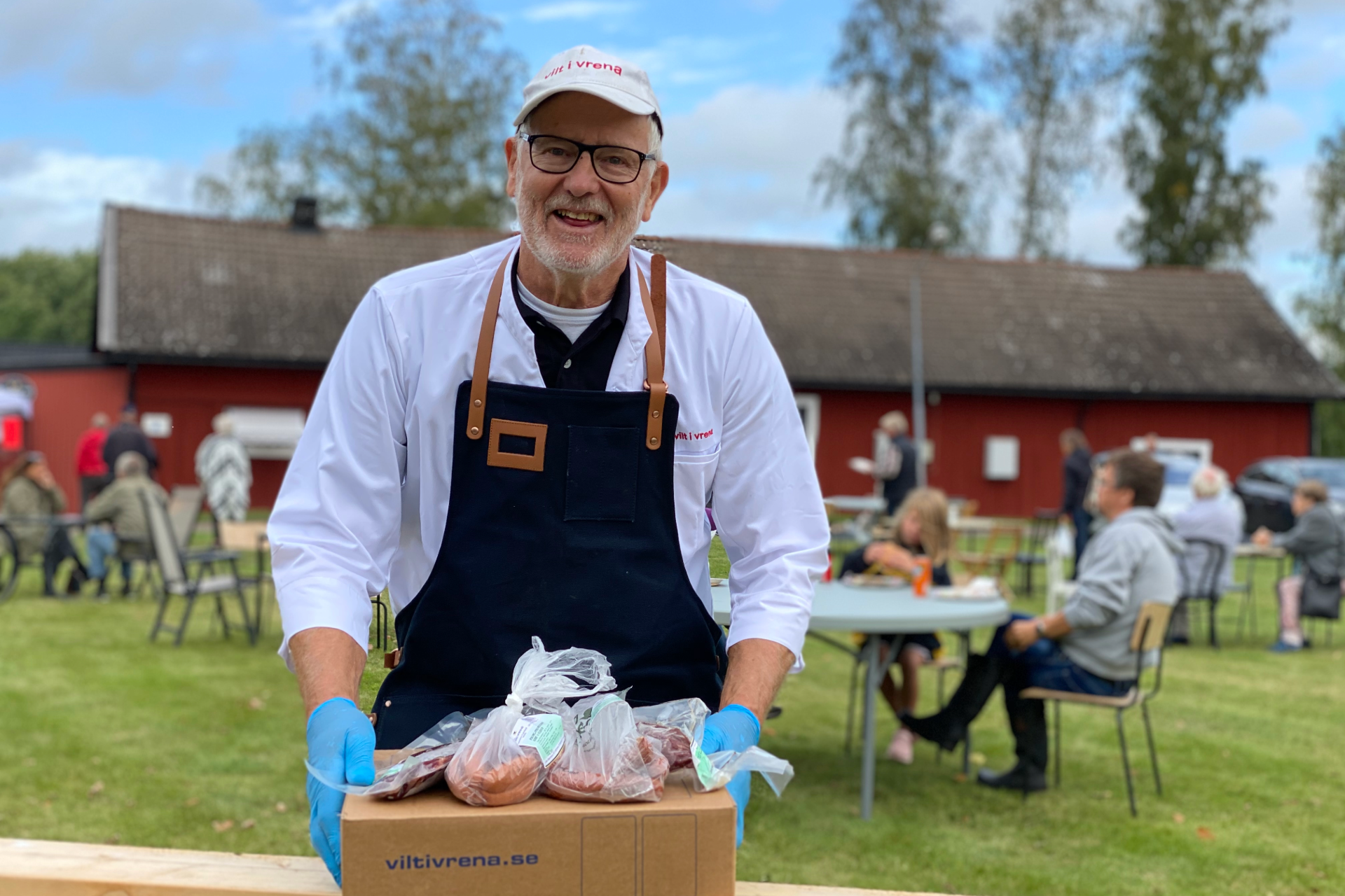 En leende man i vit skjorta och mörkblått förkläde håller en låda med vakuumförpackat kött. Han står utomhus framför ett rött hus, med folk som sitter vid bord i bakgrunden.