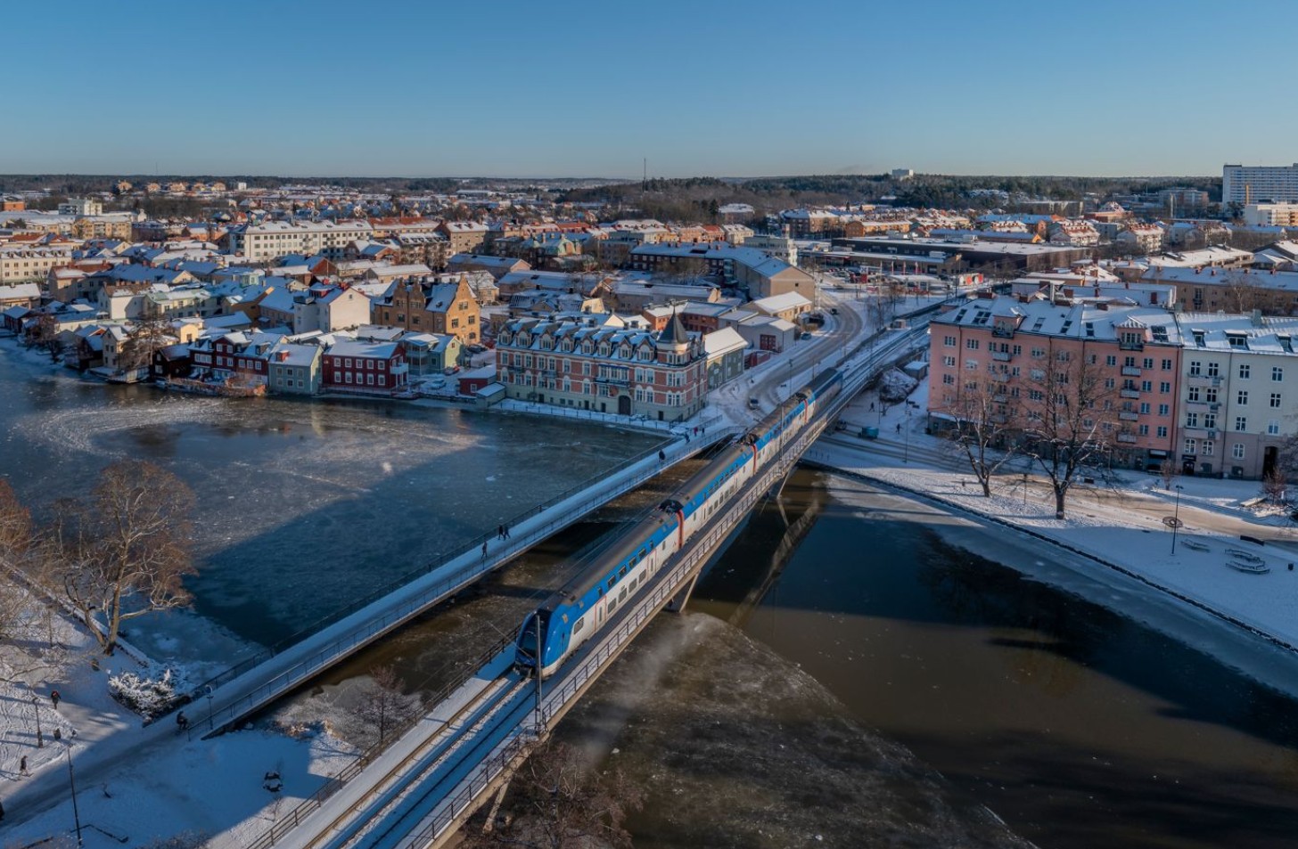 En flygbild på ett tåg som kör över en bro omgiven av vintrig stadsmiljö. 