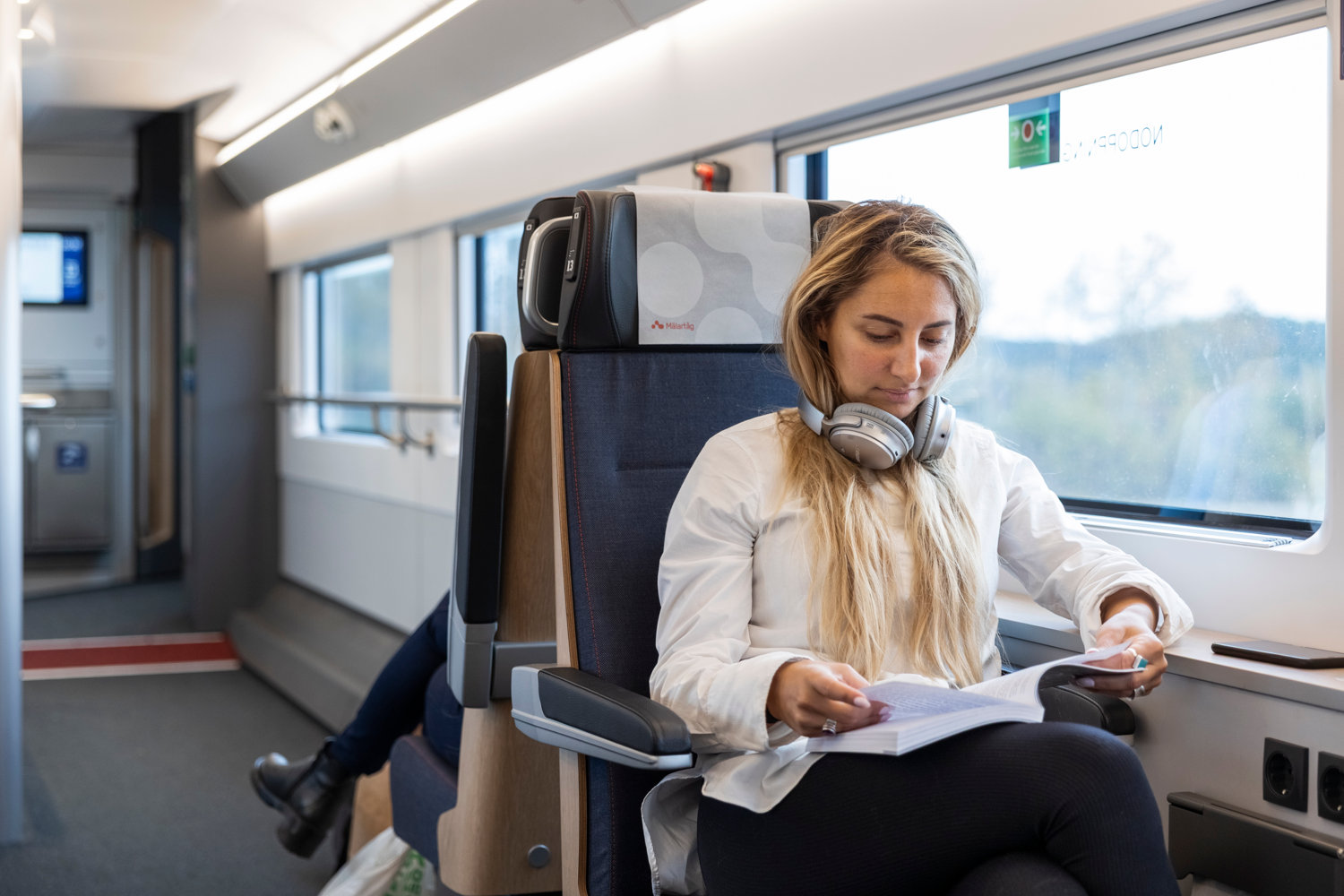 Ung kvinna som sitter och läser på ett Mälartåg med hörlurar runt halsen.