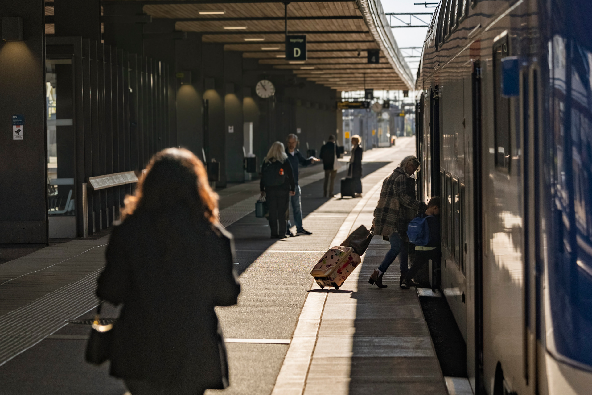 En tågstation där flera personer står på perrongen. En person med en rullväska kliver ombord på ett tåg som står vid plattformen. Andra personer väntar eller går längs perrongen.