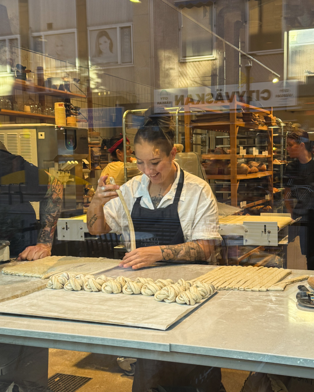 Kvinna som bakar bullar och ler bakom ett skyltfönster