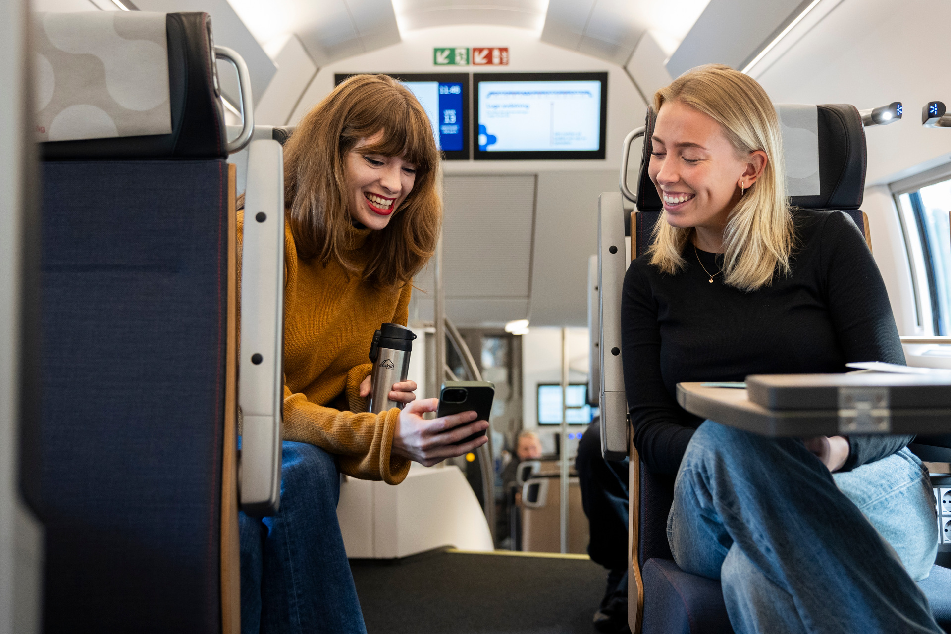 Two young women sit in a train compartment, laughing while looking at a mobile phone. One is holding a thermos, and the train appears modern with information screens in the background.