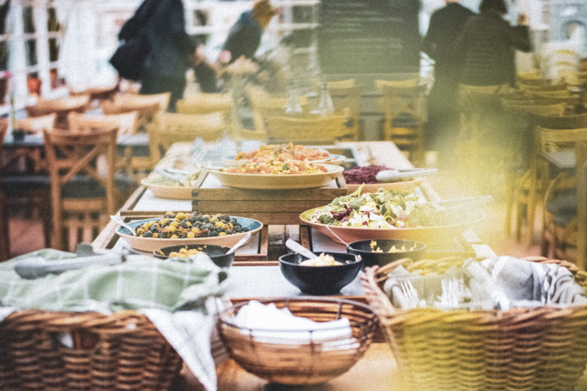 Ett buffébord med sallader, grönsaker och små skålar, omgiven av trästolar i en ljus restaurangmiljö.